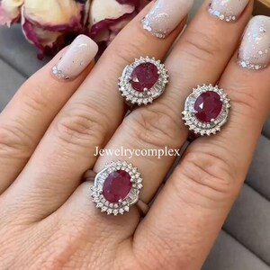 Puede incluir: Un conjunto de joyería compuesto por dos anillos y un pendiente, cada uno con un rubí rojo intenso de corte ovalado rodeado por un halo de pequeños diamantes blancos en metal plateado. El texto Jewelrycomplex es visible en el centro.