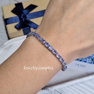 Può includere: Un bracciale tennis con una fila continua di pietre preziose color viola chiaro dal taglio ovale, incastonate in una fascia di metallo color argento. Il bracciale è indossato al polso e il testo Jewelrycomplex è visibile nell'immagine.