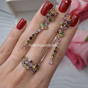 May include: A jewelry set featuring a ring and dangling earrings. The ring and earrings are adorned with a cluster of colorful gemstones in shades of pink, yellow, green, and brown. The jewelry is set against a soft background.