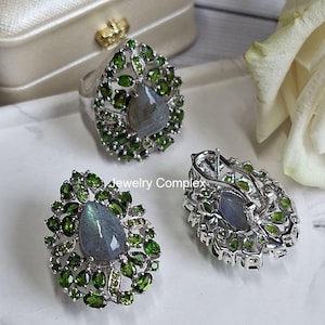 May include: A jewelry set featuring a ring and earrings. The ring and earrings have a teardrop-shaped design with a central gray stone surrounded by green gemstones. The jewelry is set on a white surface with a white rose in the background.