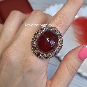 Puede incluir: Un llamativo anillo con una gran piedra preciosa ovalada de color rojo intenso, rodeada por un halo de piedras más pequeñas y coloridas. El anillo tiene una banda plateada. El texto "Jewelrycomplex" es visible.