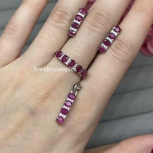 Puede incluir: Un conjunto de joyas con un anillo, pendientes y un colgante. Cada pieza está adornada con piedras preciosas rojas oscuras de corte ovalado y pequeñas piedras de acento transparentes. Las joyas se exhiben sobre un fondo claro, resaltando los colores vibrantes y el diseño intrincado.