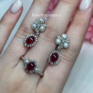 Puede incluir: Un conjunto de joyas con un anillo y pendientes. El anillo y los pendientes tienen un engaste de metal plateado con una piedra roja ovalada central, rodeada de pequeñas piedras transparentes y perlas. Los pendientes son colgantes. El texto "Jewelrycomplex" es visible.
