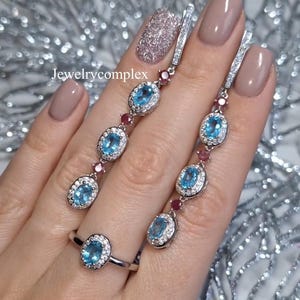Può includere: Un set di gioielli in argento con un anello e orecchini pendenti. Ogni pezzo presenta pietre preziose ovali blu chiaro circondate da piccole pietre trasparenti. Gli orecchini e l'anello sono impreziositi da piccole pietre preziose rosse. Il testo "Jewelrycomplex" è visibile.