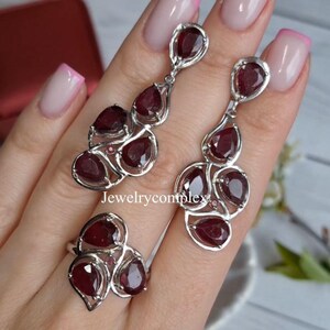 May include: A jewelry set featuring a ring and a pair of earrings. Each piece has a silver-colored metal setting and is adorned with multiple teardrop-shaped, deep red gemstones. The earrings are long and dangle, while the ring has a cluster of stones.