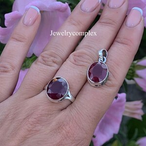 Op de afbeelding: Een zilveren ring en een bijpassende zilveren hanger, elk voorzien van een gefacetteerde ovale dieprode edelsteen. De sieradenset wordt getoond met de tekst Jewelrycomplex zichtbaar in het midden. De stukken hebben gepolijste metalen zettingen.