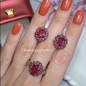 Op de afbeelding: Een sieradenset met een ring en oorbellen met een bloemmotief. Elk stuk heeft een grote, ovaalvormige, dieprode edelsteen omringd door kleinere roze edelstenen. De tekst "Jewelrycomplex" is zichtbaar.
