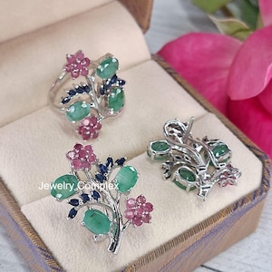 Op de afbeelding: Een sieradenset met een ring, oorbellen en een broche, allemaal met een bloemmotief. De stukken zijn zilverkleurig en versierd met ovaal geslepen groene edelstenen, roze bloemaccenten en kleine blauwe accenten. De set wordt gepresenteerd in een sieradendoosje.