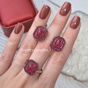 Op de afbeelding: Een driedelige sieradenset met een ring en een paar oorbellen. Elk stuk heeft een centrale, grote, rode edelsteen omringd door kleinere rode stenen. De sieraden zijn geplaatst tegen een neutrale achtergrond, met de tekst "Jewelrycomplex" zichtbaar.