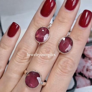 Peut inclure: Un ensemble de bijoux comprenant une bague et deux boucles d'oreilles pendantes, tous ornés de pierres précieuses rouge foncé de taille ovale. La bague est portée au doigt, et les boucles d'oreilles sont présentées. Le texte "Jewelrycomplex" est visible.