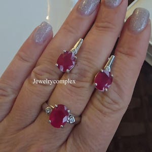 May include: A jewellery set consisting of a ring and two matching earrings, each featuring a faceted oval-cut deep red gemstone set in silver-toned metal with small clear accent stones. The text Jewelrycomplex is visible on the image.