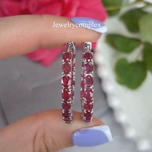 May include: Silver-toned hoop earrings, each featuring a line of oval, deep red gemstones. The earrings are held up, showcasing the design. The text "Jewelrycomplex" is visible in the image.