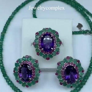 Op de afbeelding: Een sieradenset bestaande uit een ketting, een ring en twee oorbellen. Elk stuk bevat een centrale ovale paarse amethist omringd door een halo van kleine roze robijnen en groene smaragden. De tekst Jewelrycomplex is in het midden zichtbaar.