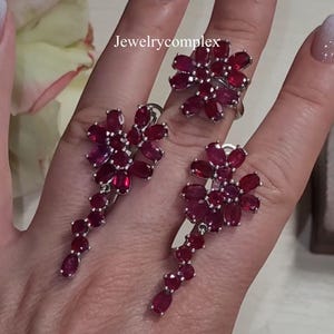 Puede incluir: Un conjunto de joyería compuesto por un anillo y dos pendientes colgantes, cada uno con racimos de piedras preciosas ovaladas de color rojo intenso dispuestas en motivos florales. Los engastes son plateados. El texto Jewelrycomplex es visible.
