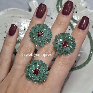 Puede incluir: Un conjunto de joyas compuesto por un anillo y dos pendientes. Cada pieza presenta un diseño floral con piedras preciosas verdes y rojas. Las joyas se exhiben en una mano con esmalte de uñas burdeos. El texto "Jewelrycomplex" es visible.