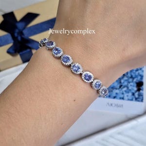 Può includere: Un bracciale in argento con pietre preziose ovali blu chiaro, circondate da pietre più piccole e trasparenti. Il bracciale è indossato su un polso, con il testo "Jewelrycomplex" sullo sfondo.