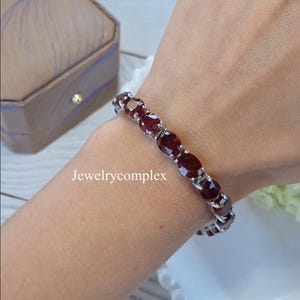 Puede incluir: Un brazalete de plata con piedras preciosas rojas oscuras de corte ovalado. El brazalete se muestra en una muñeca, destacando el diseño y el color de la joya. Incluye el texto "Jewelrycomplex".