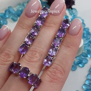Puede incluir: Un conjunto de joyas compuesto por dos pendientes largos y un anillo, cada uno con una fila vertical de piedras preciosas amatista moradas de corte ovalado engastadas en metal plateado. El texto Jewelrycomplex es visible en la imagen.