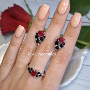 Pode incluir: Um conjunto de anel e brincos de prata. O anel e os brincos apresentam um design de aglomerado com uma pedra preciosa vermelha central rodeada por pedras pretas de corte quadrado. As joias são exibidas em uma mão com esmalte de unhas neutro.