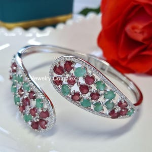 May include: Silver-tone open cuff bracelet. The bracelet is adorned with alternating red and green gemstones, framed by a border of small, clear stones. The jewellery is presented on a white surface.