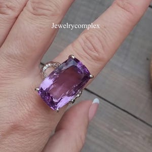 Pode incluir: Um anel de prata com uma grande pedra preciosa ametista retangular facetada, presa por quatro garras. A banda tem um design decorativo de metal vazado. O texto Jewelrycomplex é visível acima do anel na superfície da imagem.