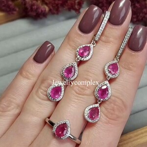 Puede incluir: Un par de pendientes largos de plata y un anillo a juego, cada uno con varias piedras preciosas rosas en forma de pera rodeadas de pequeñas piedras transparentes. El conjunto de joyería muestra un diseño consistente con piedras rosas.