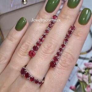 Puede incluir: Un anillo de plata y dos pendientes a juego, cada uno con una hilera de piedras preciosas redondas de color rojo intenso. El conjunto de joyas se muestra con el texto Jewelrycomplex visible en el centro sobre un marco de metal plateado.
