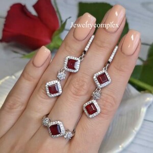 Op de afbeelding: Een sieradenset met een ring en oorhangers. Elk stuk heeft een vierkante, dieprode edelsteen omringd door kleine, heldere stenen. De oorbellen en ring zijn zilverkleurig. De tekst "jewelrycomplex" is zichtbaar in de afbeelding.