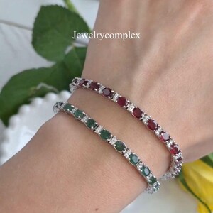 Puede incluir: Se muestran dos pulseras de tenis, una con piedras de color verde esmeralda y otra con piedras de color rojo rubí. Cada pulsera presenta una fila de piedras preciosas de talla ovalada intercaladas con pequeñas piedras transparentes. Las pulseras están en una muñeca.