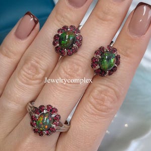 May include: A jewelry set featuring a ring and earrings. Each piece has a dark opal center stone surrounded by small, red gemstones. The ring and earrings are set in a silver metal. The text "Jewelrycomplex" is visible.