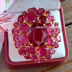 May include: A statement ring with a gold setting, adorned with numerous oval and round-cut rubies. The central ruby is a large oval shape, encircled by smaller rubies. The ring is presented in a red jewellery box. The text "Jewelry Complex" is visible.