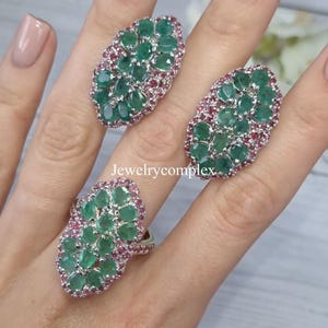 Op de afbeelding: Drie ovale ringen met clusters van smaragdgroene stenen en kleinere roze edelstenen. De ringen worden getoond op een licht oppervlak. De tekst "jewelrycomplex" is zichtbaar.