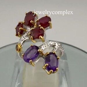 Op de afbeelding: Een zilveren ring met een bloemmotief, met ovale rode en paarse edelstenen. De ring heeft gouden accenten en de tekst "jewelrycomplex". De edelstenen zijn in een clusteropstelling geplaatst.