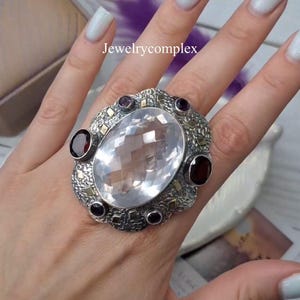 Pode incluir: Um anel ornamentado com uma grande pedra preciosa clara e facetada no centro. O anel tem uma banda prateada texturizada, acentuada com pedras preciosas menores de cor vermelho escuro e roxo. O texto "Jewelrycomplex" é visível.