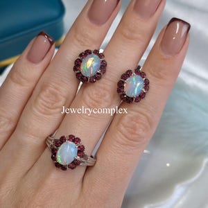 Könnte beinhalten: Ein Schmuckset mit einem Opalring und passenden Ohrringen. Jedes Schmuckstück hat einen ovalen Opal in der Mitte, umgeben von kleinen, runden, tiefroten Edelsteinen. Der Ring und die Ohrringe sind aus silberfarbenem Metall gefertigt. Der Text "Jewelrycomplex" ist sichtbar.