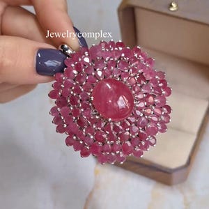 Op de afbeelding: Een ronde hanger met een grote, gladde, ovale roze edelsteen in het midden, omringd door meerdere lagen kleinere, gefacetteerde, ronde en ovale roze edelstenen gezet in zilverkleurig metaal. De tekst Jewelrycomplex is zichtbaar.