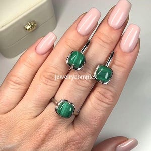 Pode incluir: Um conjunto de anel e brincos de prata com pedras de malaquite verde quadrada. As joias são exibidas em uma mão com esmalte de unhas rosa. O texto "jewelrycomplex" é visível.