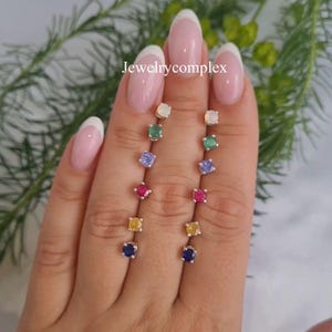 May include: A close-up of two rows of square-cut gemstone stud earrings in various colors, including opal, emerald, amethyst, ruby, yellow sapphire, and sapphire. The earrings are set in silver. The text "Jewelrycomplex" is visible.