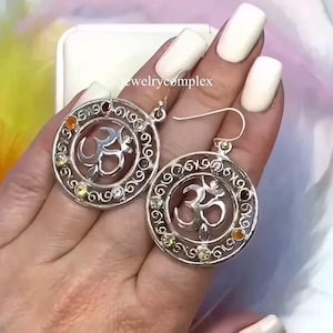 May include: Silver earrings featuring the Om symbol, encircled by gemstones and a decorative filigree design. The round earrings have a hook fastening. The text "jewelrycomplex" is visible.