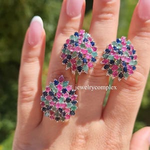 May include: A set of three gemstone rings with a cluster design. Each ring features a silver setting and is adorned with a mix of pink, blue, and green gemstones. The rings are displayed on a hand with a blurred green background.