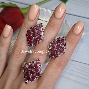 Op de afbeelding: Een set van drie zilveren ringen, elk versierd met talrijke ovale, dieprode edelstenen. De ringen zijn gerangschikt op een lichtgekleurd oppervlak, dat het ingewikkelde ontwerp en de levendige kleur van de sieraden benadrukt. De tekst "jewelrycomplex" is zichtbaar.