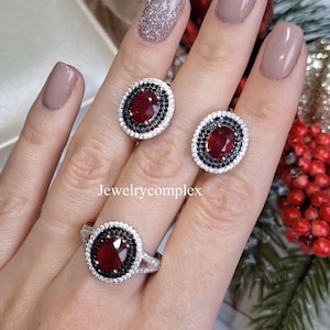 Può includere: Un set di gioielli con anello e orecchini abbinati. Ogni pezzo ha una grande pietra ovale rosso scuro circondata da un alone di pietre nere e bianche. Il testo "Jewelrycomplex" è visibile.