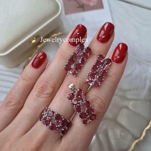Op de afbeelding: Een sieradenset met een ring, hanger en oorbellen, allemaal versierd met levendige rode edelstenen. De zilverkleurige metalen zetting complementeert de rijke kleur van de stenen. De tekst "Jewelrycomplex" is zichtbaar.
