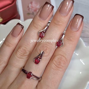 May include: A silver jewellery set comprising a ring and earrings, each featuring a large, oval, red gemstone. The earrings and ring are accented with smaller red stones. The text "jewelrycomplex" is visible.