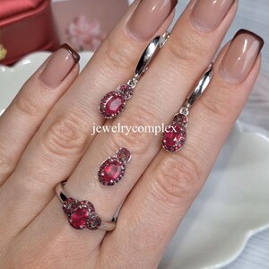 May include: A jewelry set featuring a ring and earrings with oval-shaped, ruby-red gemstones. The ring has a cluster of three stones, while the earrings and pendant have a single stone surrounded by smaller gems. The jewelry is silver-toned.