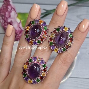 Puede incluir: Tres anillos de piedras preciosas con un amatista ovalado central rodeado por un halo de gemas coloridas. Los anillos se muestran en una mano con uñas cuidadas. El texto "Jewelrycomplex" es visible.