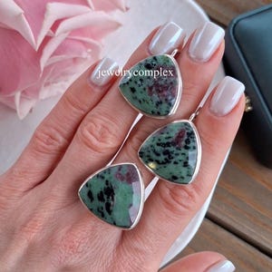 Könnte beinhalten: Drei dreieckige Anhänger mit silbernem Rand, mit grün und kastanienbraun gesprenkelten Steinen. Die Anhänger werden auf einer weißen Oberfläche präsentiert. Der Text "jewelrycomplex" ist sichtbar. Der Hintergrund umfasst eine rosa Blume und eine dunkle Box.