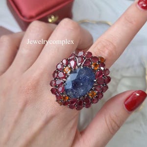 Può includere: Un anello ornato con una pietra centrale blu scuro sfaccettata, circondata da pietre più piccole rosse e arancioni. L'anello ha un design floreale ed è montato su una fascia argentata. Il testo "Jewelrycomplex" è visibile.