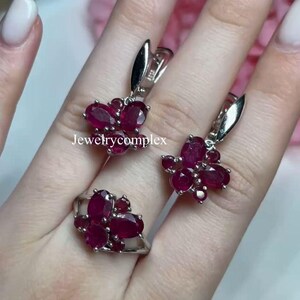 Puede incluir: Un conjunto de joyas con un anillo y pendientes, cada uno adornado con múltiples piedras preciosas rojas oscuras de corte ovalado. Los pendientes tienen un cierre plateado. El anillo y los pendientes están agrupados en un diseño floral. El texto "Jewelrycomplex" es visible.