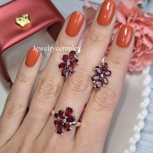 Peut inclure: Un ensemble de bijoux comprenant une bague et des boucles d'oreilles, chacun avec un motif floral. Les fleurs sont en argent et présentent des pierres précieuses rouge foncé. Le texte "jewelrycomplex" est visible.